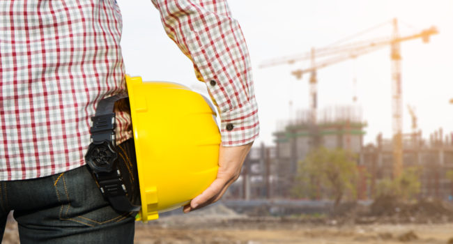 Hand's engineer worker holding yellow safety helmet with buildin Hand's engineer worker holding yellow safety helmet with building on site background.
